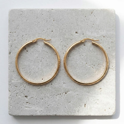 Aramat Jewels gold stainless steel hoop earrings with floral motif, 40mm diameter, on a textured stone surface.