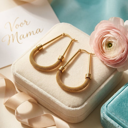 Aramat Jewels large oval gold-colored RVS hoop earrings with mesh detail, 58mm, displayed on a cream velvet box with a pink ranunculus flower.