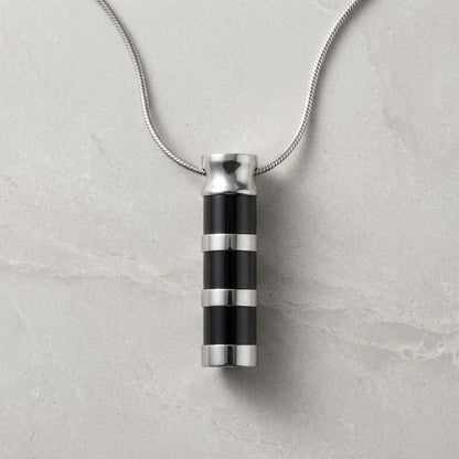 Aramat Jewels RVS cilinder ashanger met zwart-zilveren strepen, 43x11 mm. Een subtiel en tijdloos sieraad om een dierbare herinnering te bewaren.